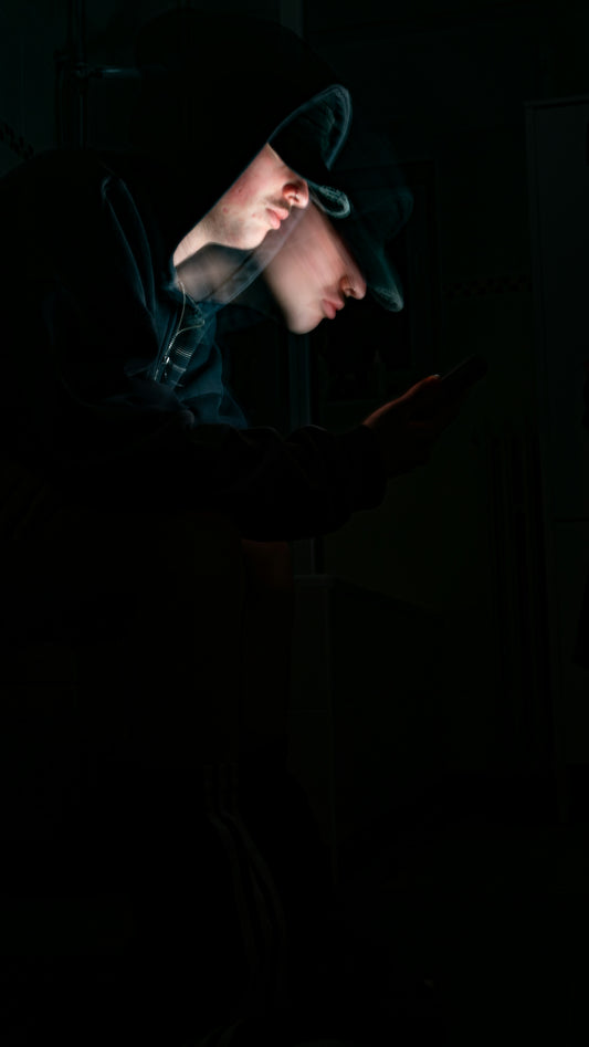 A hooded figure uses a phone in the dark.