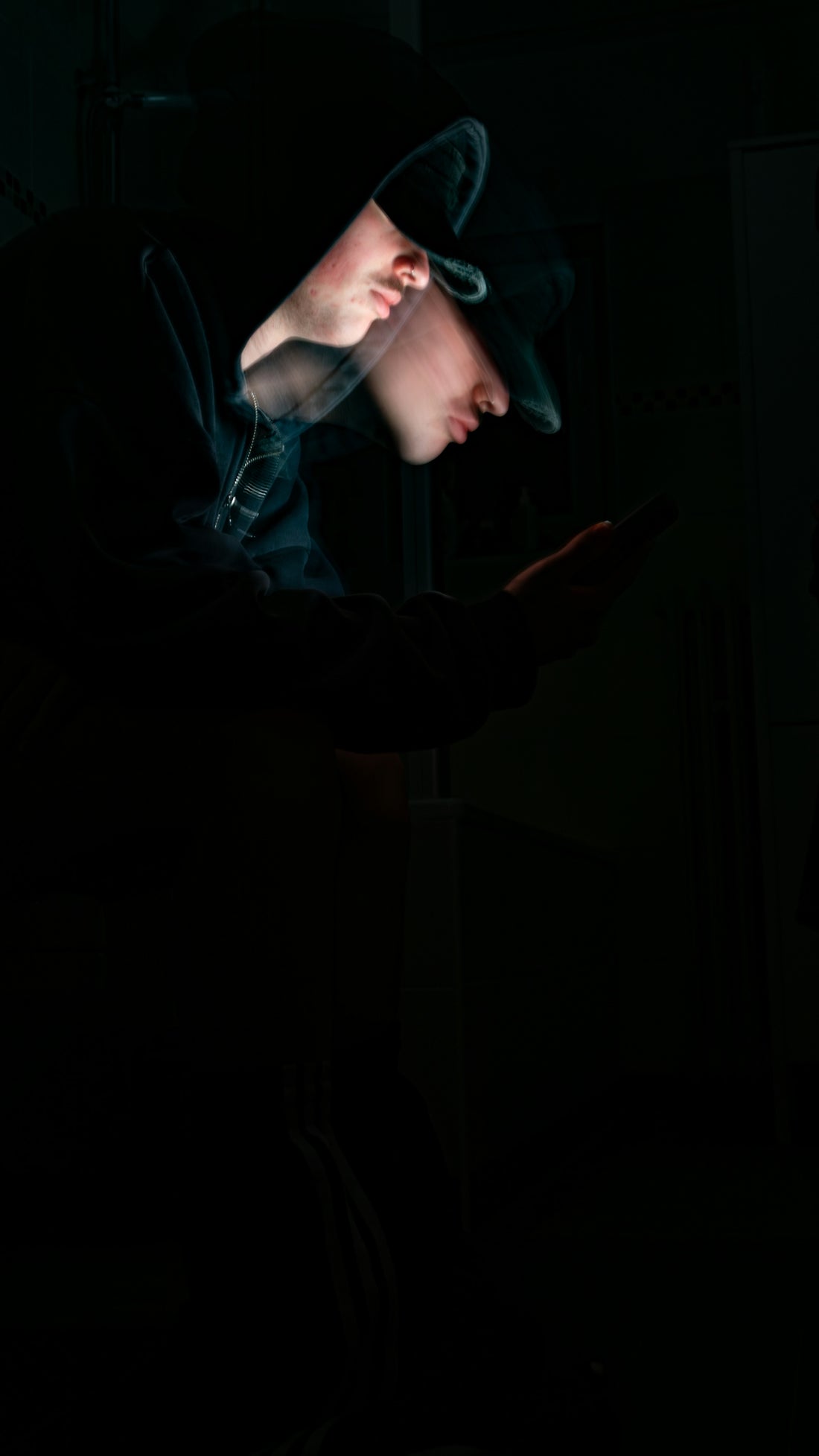 A hooded figure uses a phone in the dark.
